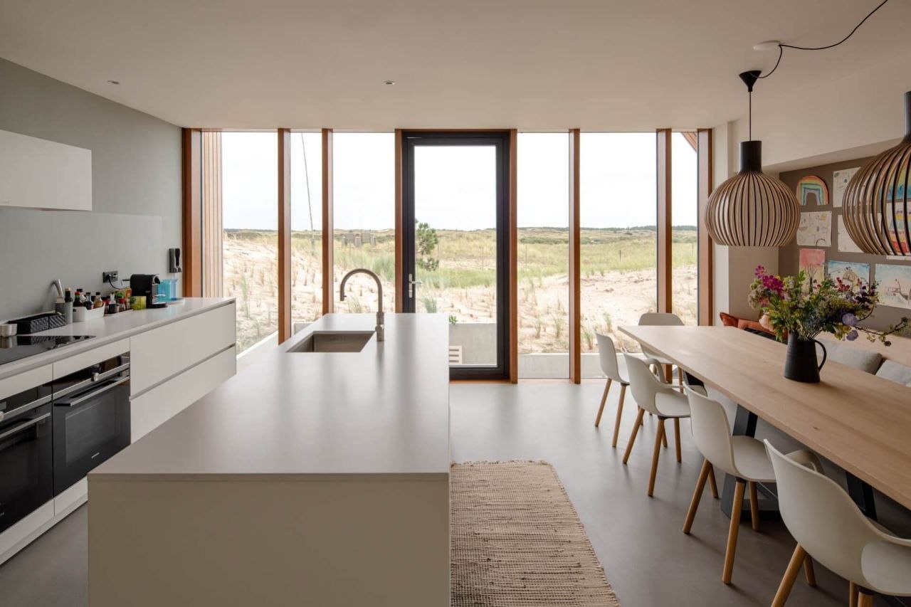 Cuisine ouverte : îlot central, grande table de bois et chaises blanches devant les baies qui donnent sur la dune