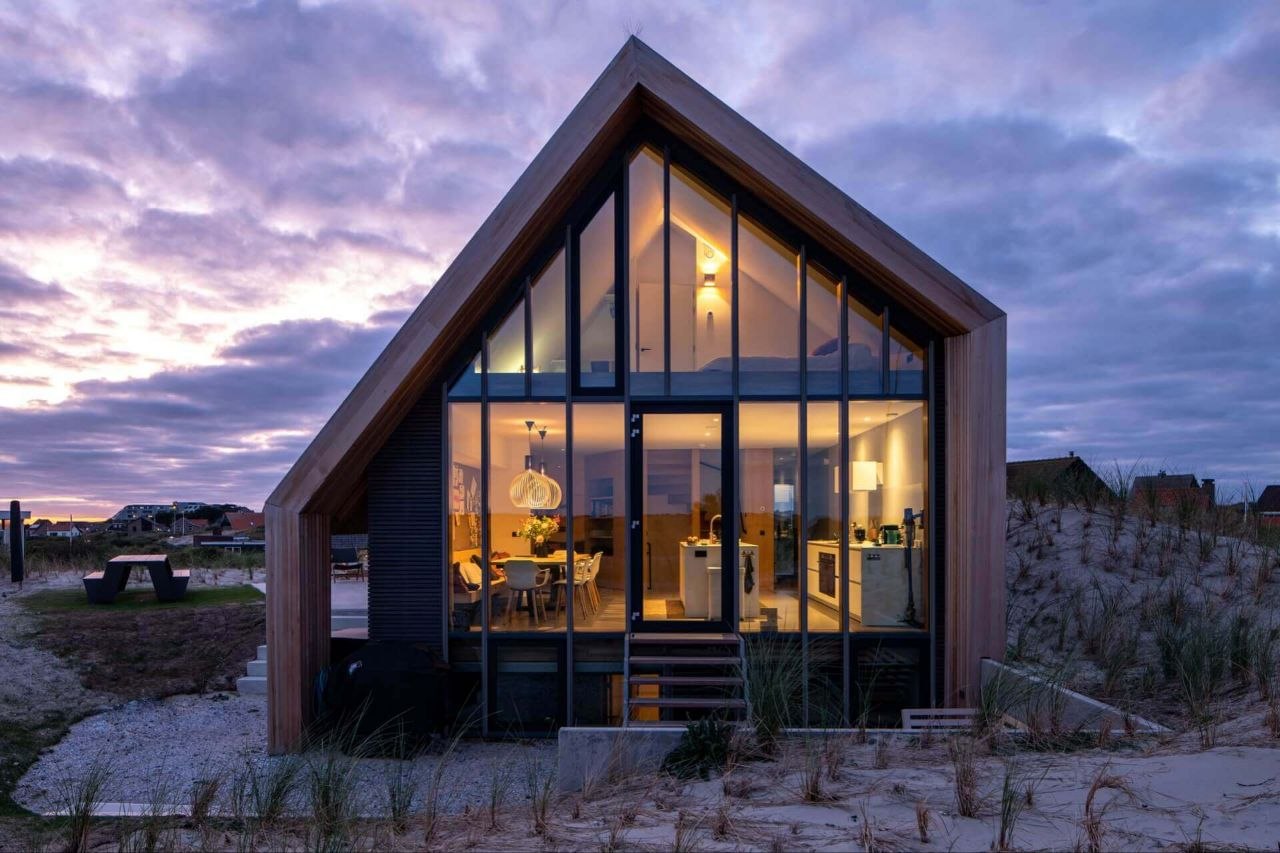 La façade vitrée de la maison allumée au crépuscule, sous un ciel violet, avec la table de pique-nique à gauche