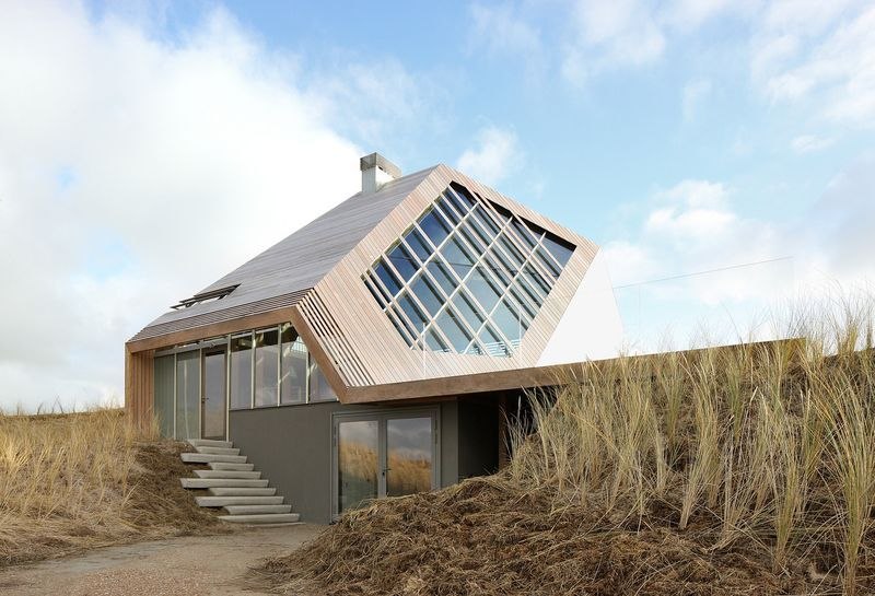 La maison en plein jour, adossée à la dune, le soleil dans l'objectif et les oyats au premier plan