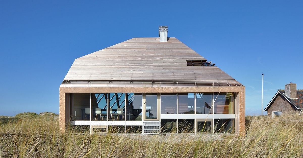 La maison dans les dunes de face : grande façade vitrée en triangle, marches de béton et oyats sur la dune, ciel bleu