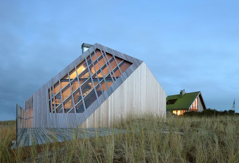 La maison au crépuscule : la façade de bois s'éclaire, la maison voisine au toit d'herbe se devine à droite