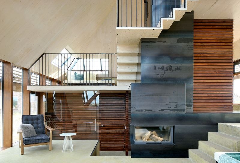 Séjour sous charpente de bois : escalier d'acier noir, cheminée, fauteuil et table d'appoint, mezzanine au-dessus