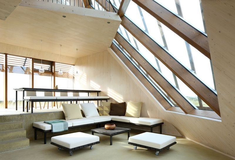 Séjour vu vers la verrière en pente : banquettes à coussins le long des vitres, table basse et poufs sur roulettes