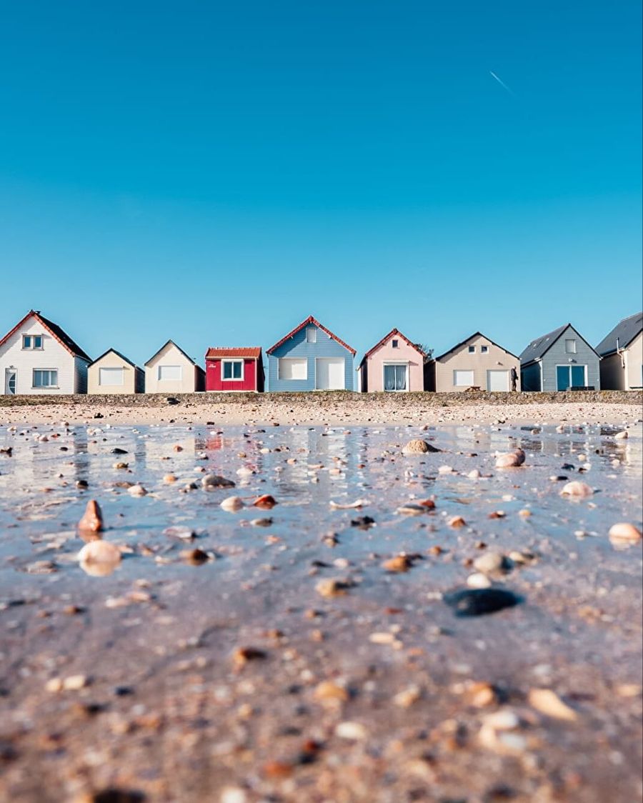 Rangée de cabanes de plage aux couleurs vives alignées au bord de l'eau, sous un ciel bleu, sable mouillé et coquillages au premier plan
